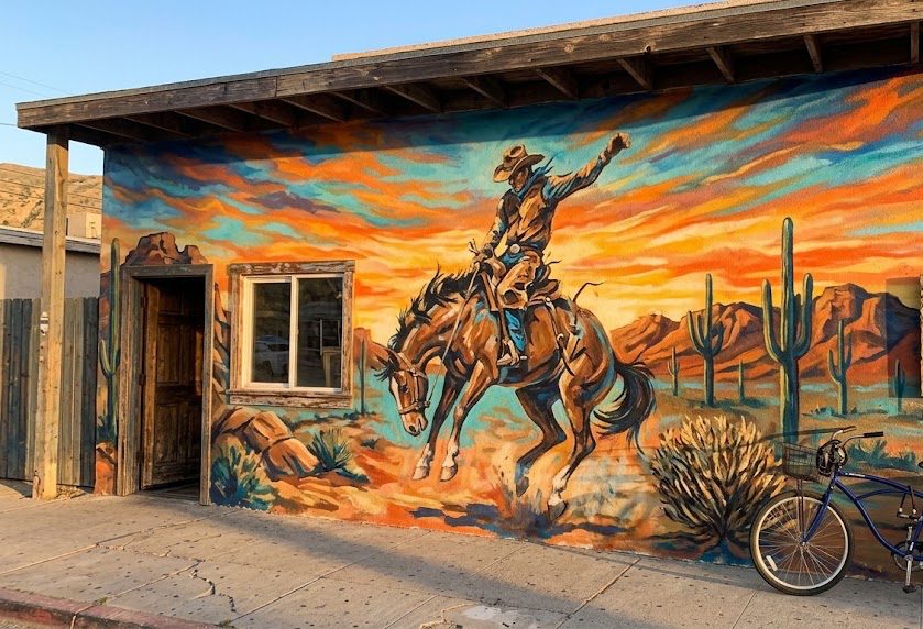 Mural of a cowboy on a bucking bronco against a desert sunset on a stucco wall with a wooden awning